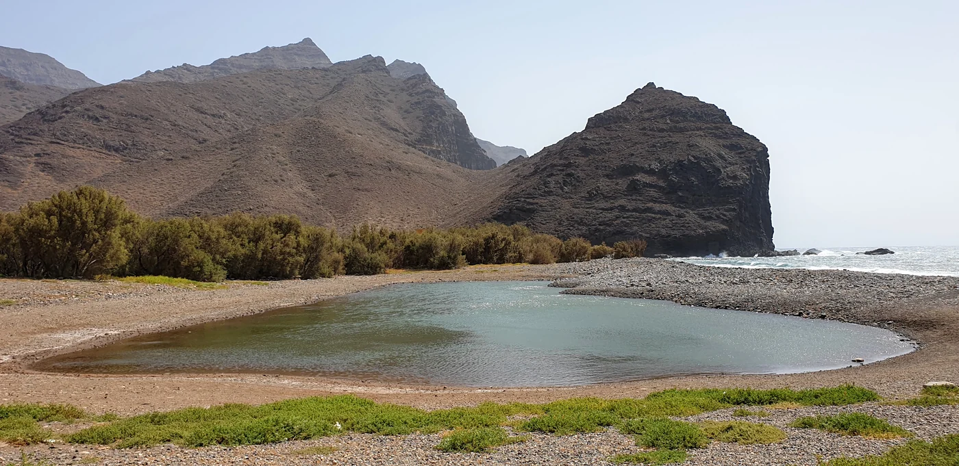 El Charco de La Aldea de San Nicolás: la laguna litoral separada del mar por una barra de callaos, con el barranco y las montañas del oeste de Gran Canaria al fondo